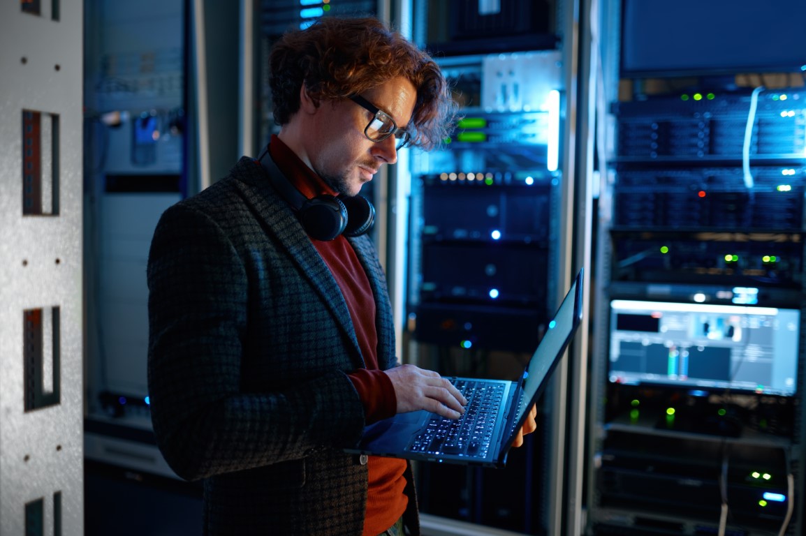 IT technician checking the servers vitals using laptop computer. Male support agent working in a dark network server room